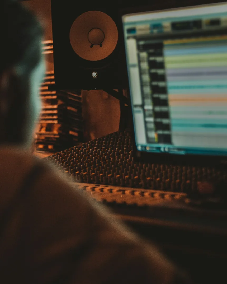 Superficie de control con faders iluminados frente a una pantalla con varias pistas de audio apiladas en la línea de tiempo